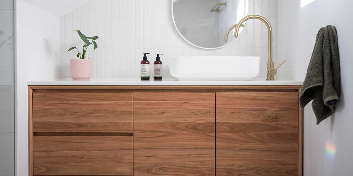 A modern wooden bathroom vanity with spacious drawers, a white vessel sink, a gold faucet, and a round mirror.